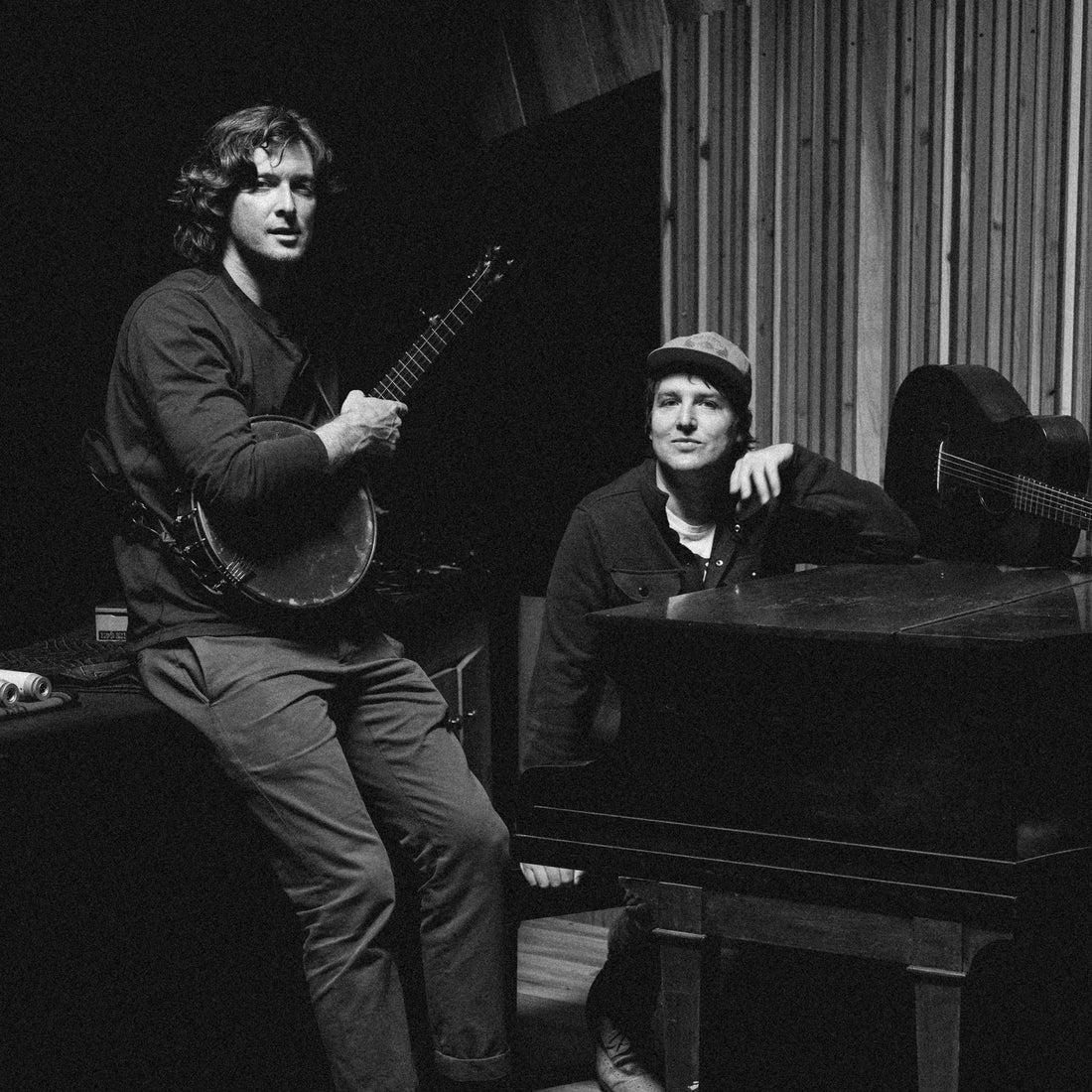 Two musicians in a recording studio, one at a piano and the other holding a banjo beside a wood-paneled wall, black-and-white portrait