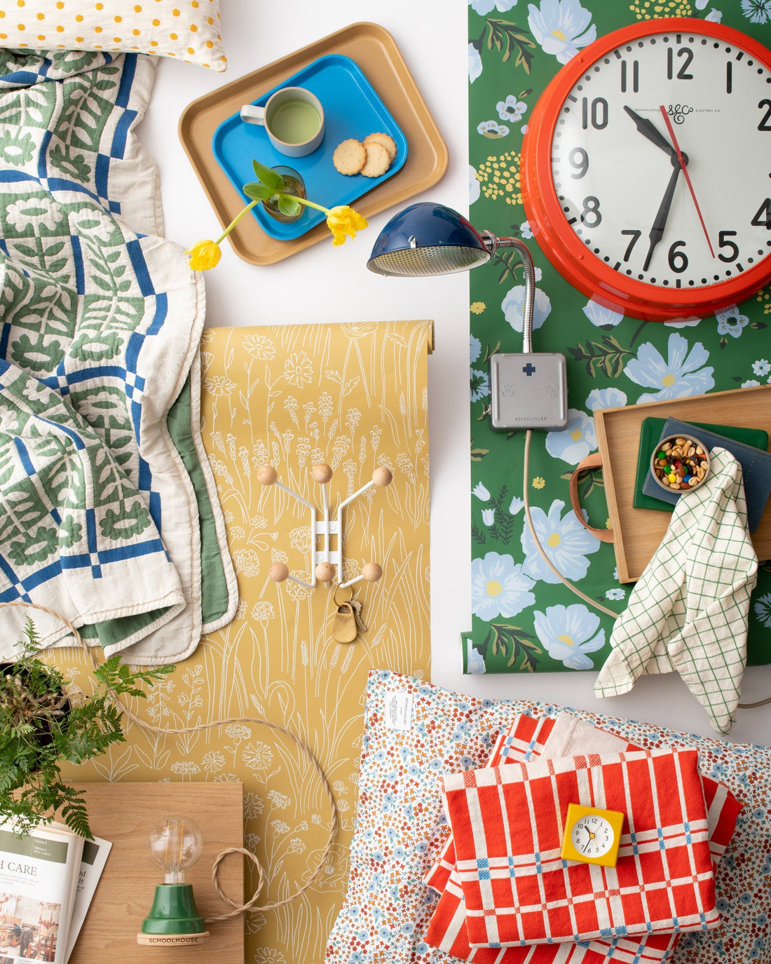 Flat lay of home goods: red wall clock, floral wallpaper roll, blue desk lamp, patterned quilts, trays with mug and snacks