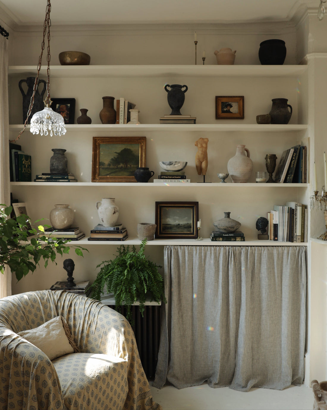 Built-in living room shelves with vintage pottery, books and framed art above a curtain, beside an armchair and fern