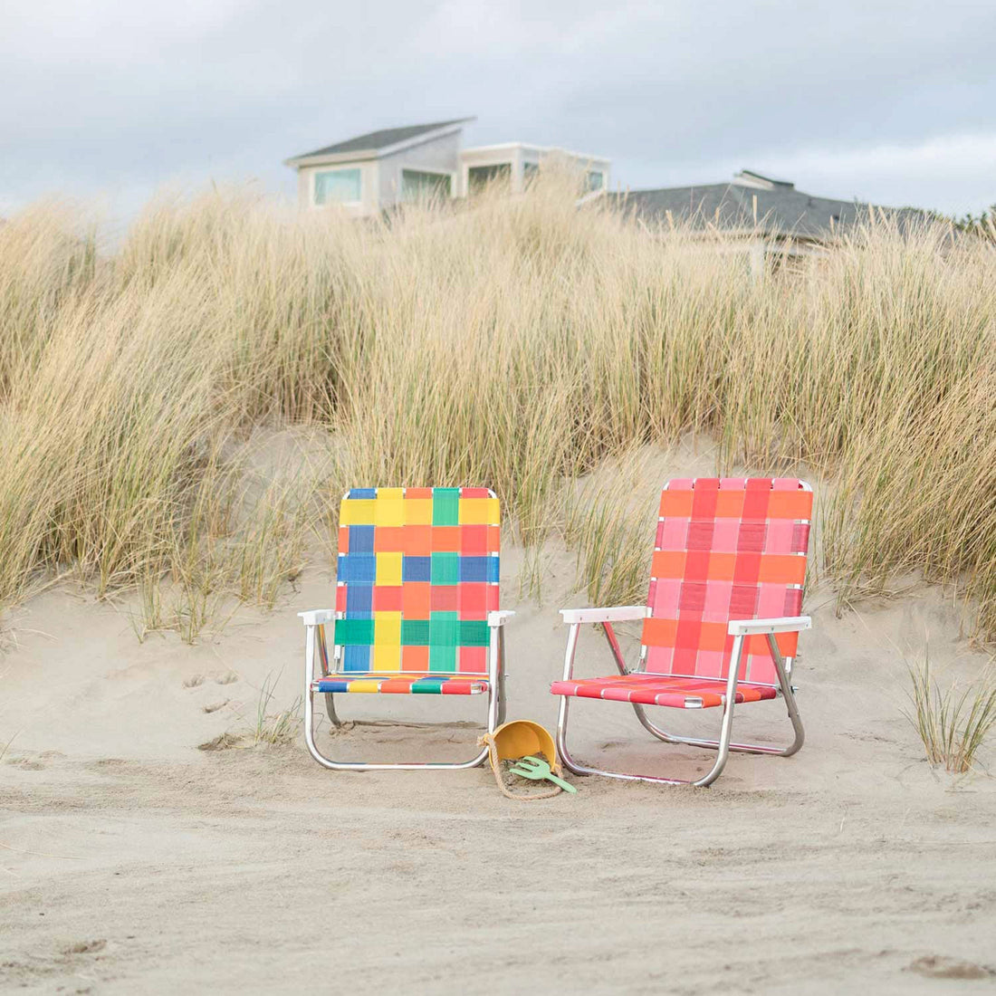 Two woven beach chairs, one multicolored and one pink, on sand in front of tall dune grasses and a distant beach house
