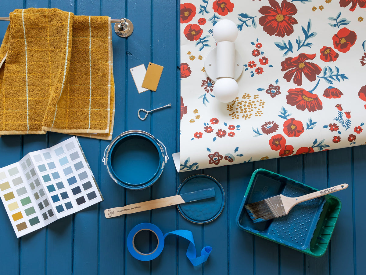 Flat lay of blue-painted wood with an open teal paint can, brush and tray, floral wallpaper sample, color swatches and a mustard towel