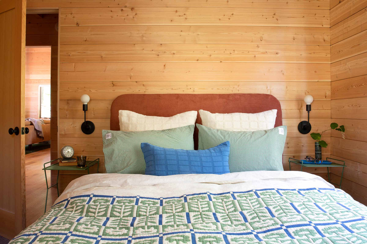 Cozy bed with rust upholstered headboard, green gingham pillows, blue lumbar pillow, and green-and-blue leaf quilt in a wood-paneled bedroom