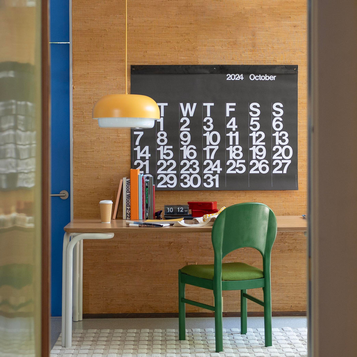 Workspace with green chair and wooden desk featuring a hanging yellow pendant and a large black wall calendar reading ‘2024 October’