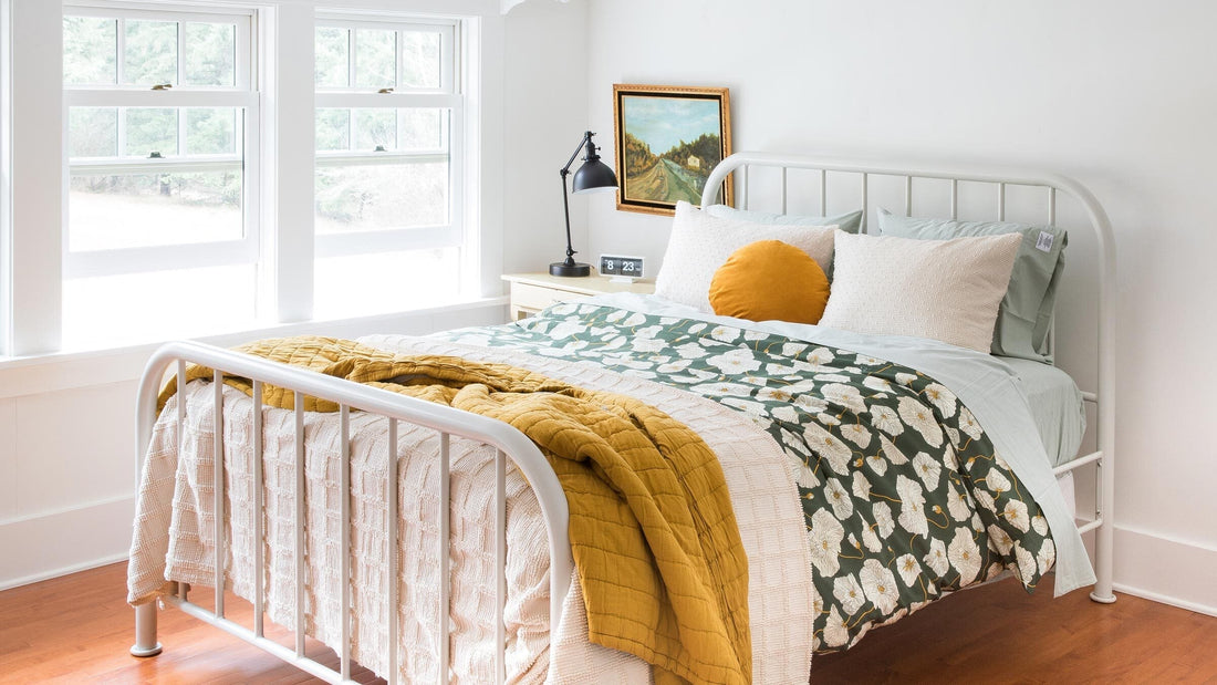 Sunlit bedroom with white metal bed dressed in green floral duvet, pale sheets, cream blanket, and mustard pillow