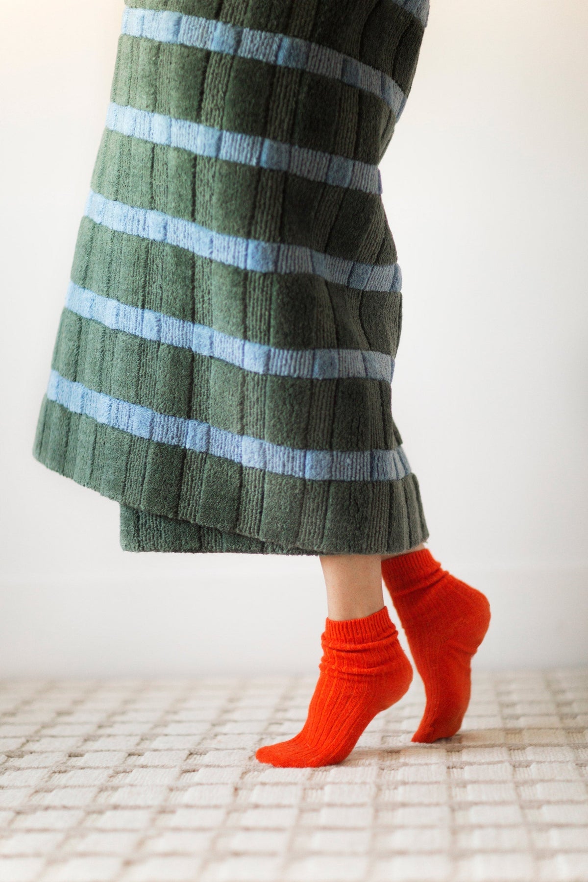 Person on tiptoes wearing bright orange ribbed socks and a green ribbed skirt with light blue horizontal stripes against a pale background