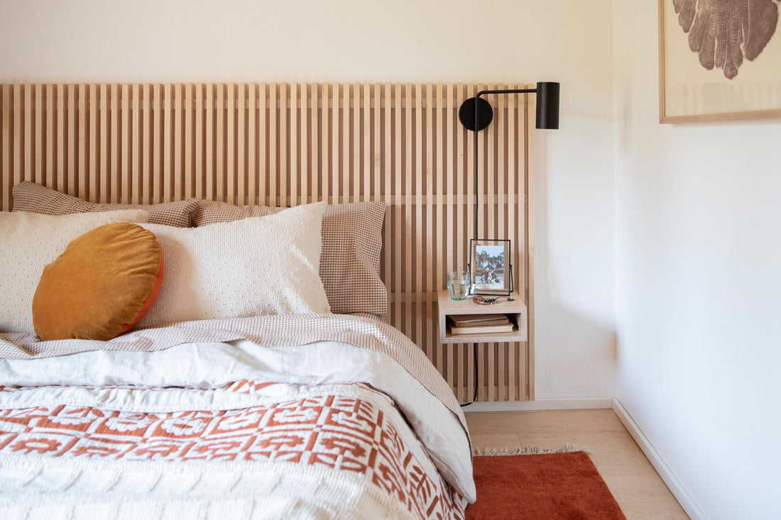 Bedroom with slatted wood headboard, cream pillows and rust patterned throw, mustard pillow, black wall sconce