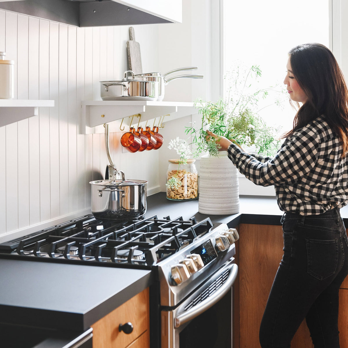 Woman arranging fresh greenery in a white ceramic vase on a bright kitchen counter beside a stainless steel gas range