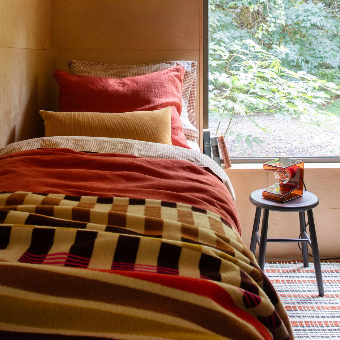 Bed styled with rust-red linen duvet, mustard lumbar pillow, gingham shams, and a checked wool throw beside a window with a leafy yard view