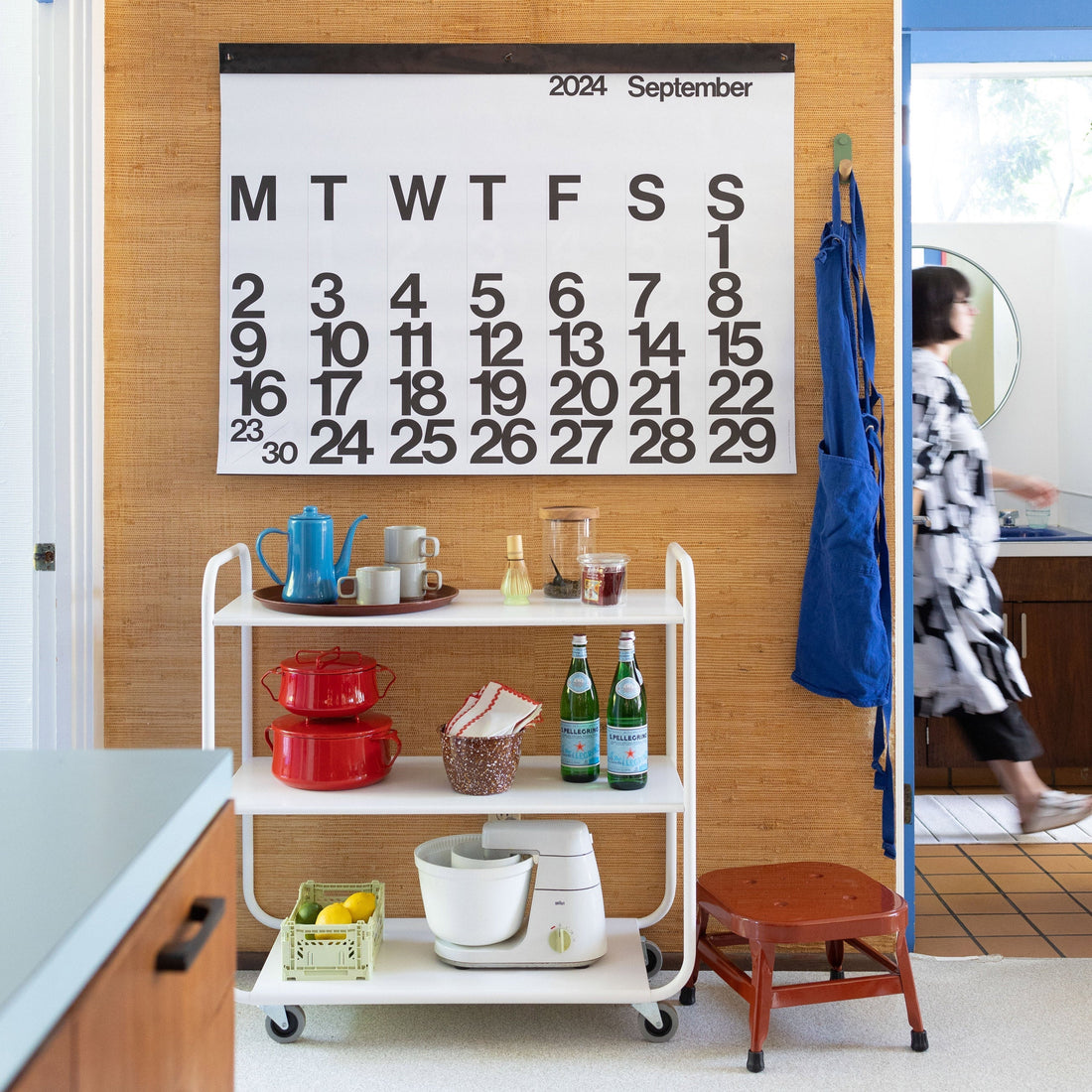 Kitchen cart neatly arranged with red pots, blue teapot, stacked mugs and two Pellegrino bottles under a large wall calendar reading ‘2024 September’, blue apron hanging and a person walking by