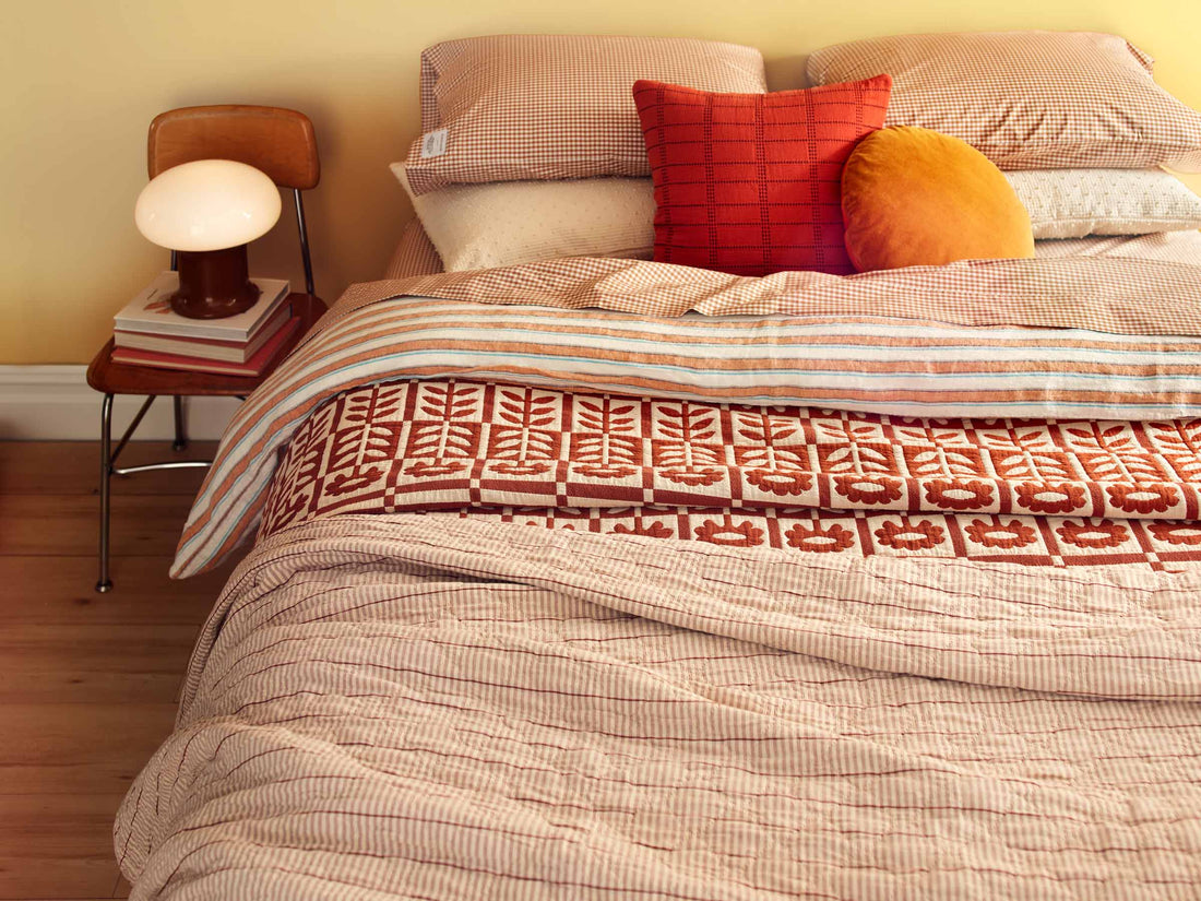 Layered bed with striped duvet, rust patterned quilt, gingham pillows, red square and round orange cushions beside a lamp