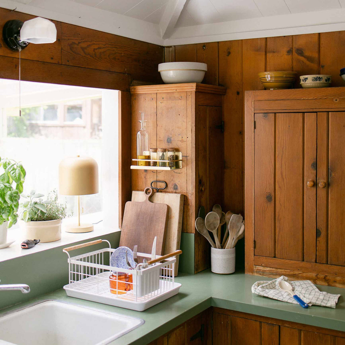 Kitchen corner with wood-paneled cabinets, a small wall sconce and yellow table lamp by a window above a green countertop with a dish rack and wooden utensils