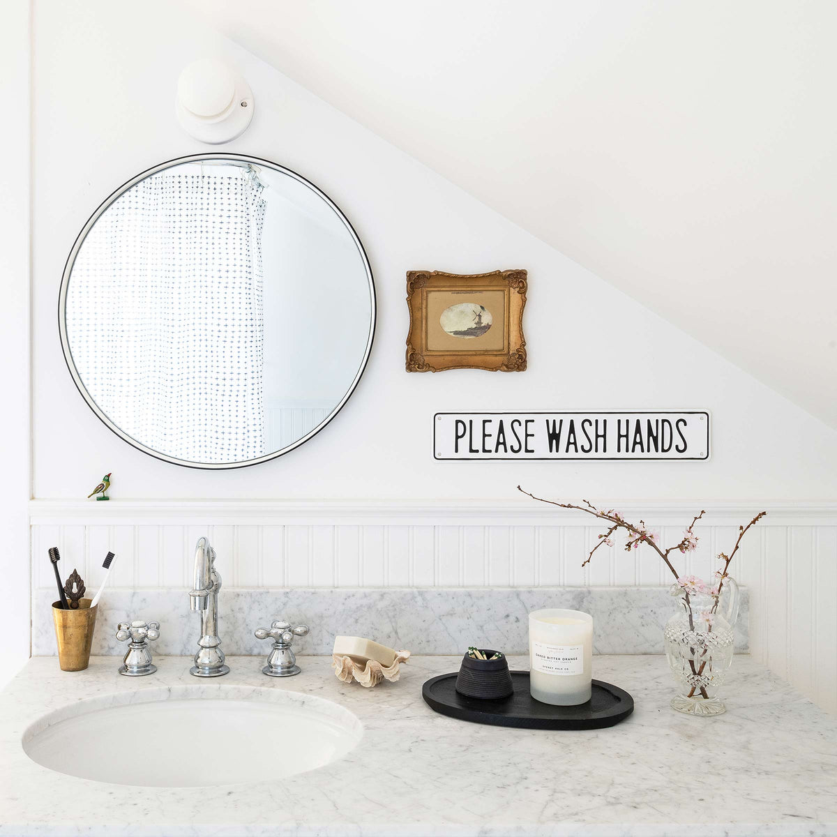 Round mirror above a marble bathroom sink with chrome faucet, small framed print, and a sign reading PLEASE WASH HANDS