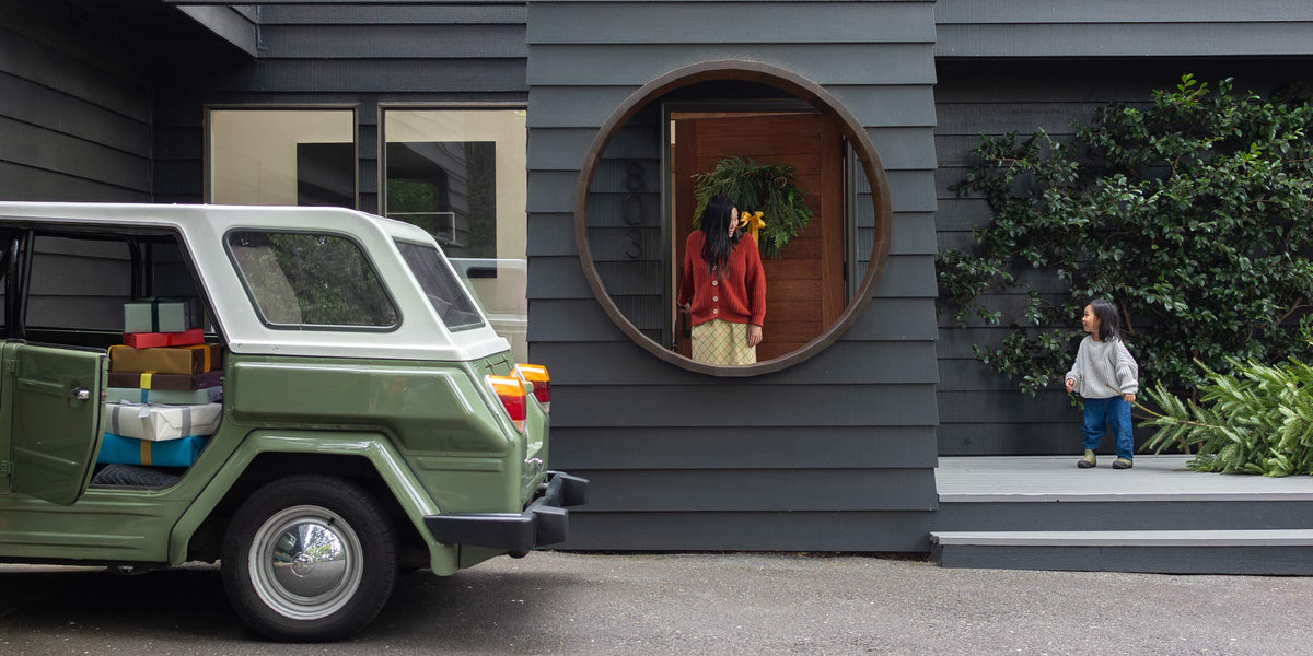 Woman in a round doorway with a holiday wreath, child on the steps, and a green SUV with wrapped gifts in the driveway