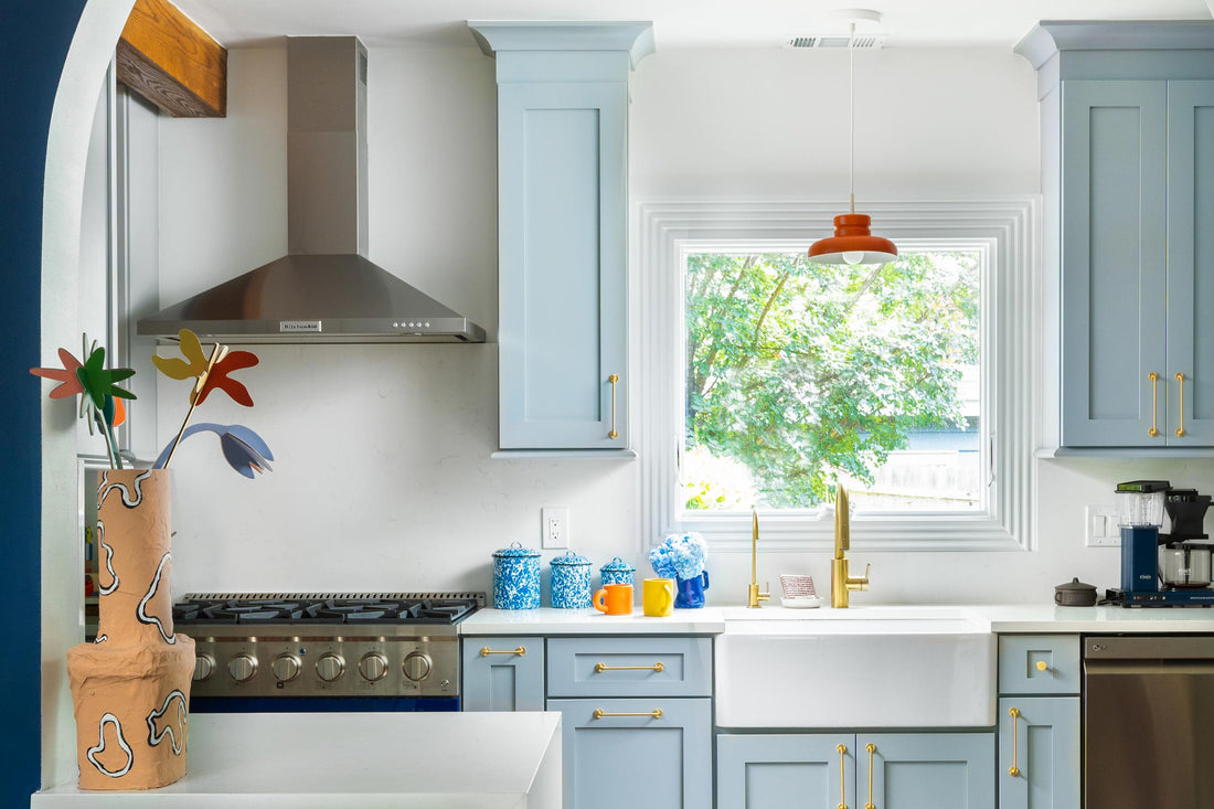 Light blue kitchen with farmhouse sink beneath a window, steel range, orange pendant and blue canisters
