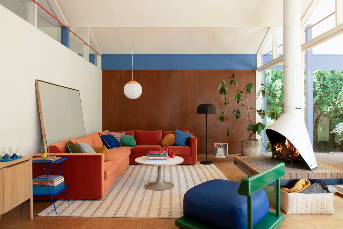 Midcentury-inspired living room with burnt-orange sectional sofa, blue ottoman, hanging globe pendant, white cone fireplace and garden-facing windows