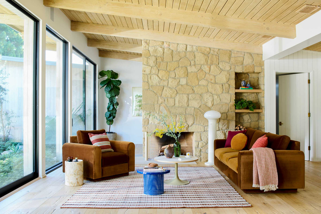 Midcentury modern living room styled with a stone fireplace, rust velvet sofa and armchair, striped rug, and floor-to-ceiling windows overlooking a garden