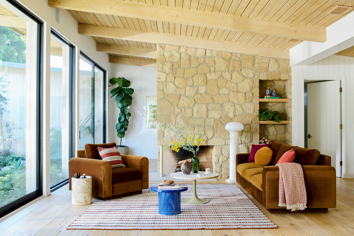 Midcentury modern living room styled with a stone fireplace, rust velvet sofa and armchair, striped rug, and floor-to-ceiling windows overlooking a garden