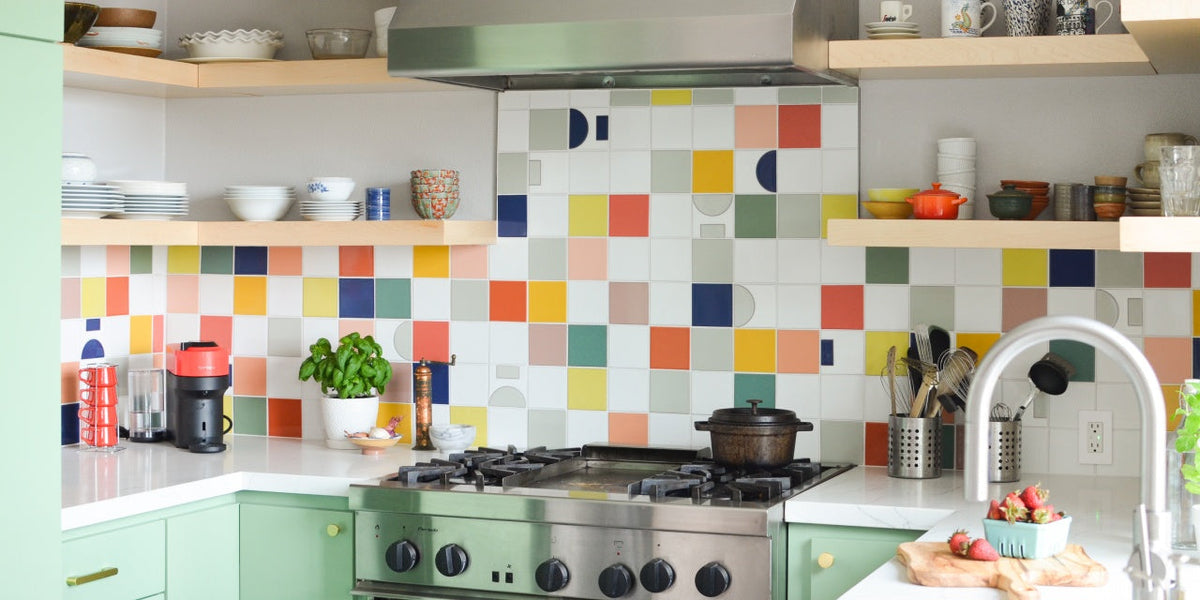 Bright kitchen with colorful geometric tile backsplash, mint-green lower cabinets, stainless gas range, open wood shelves, and countertop accessories