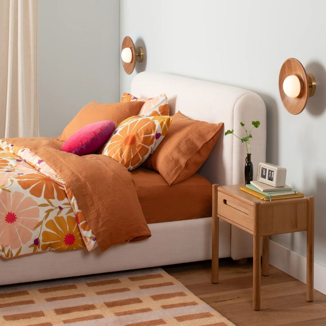 Bed with rust-orange duvet and floral cover, layered pillows and pink cushion beside an oak nightstand and wall sconces