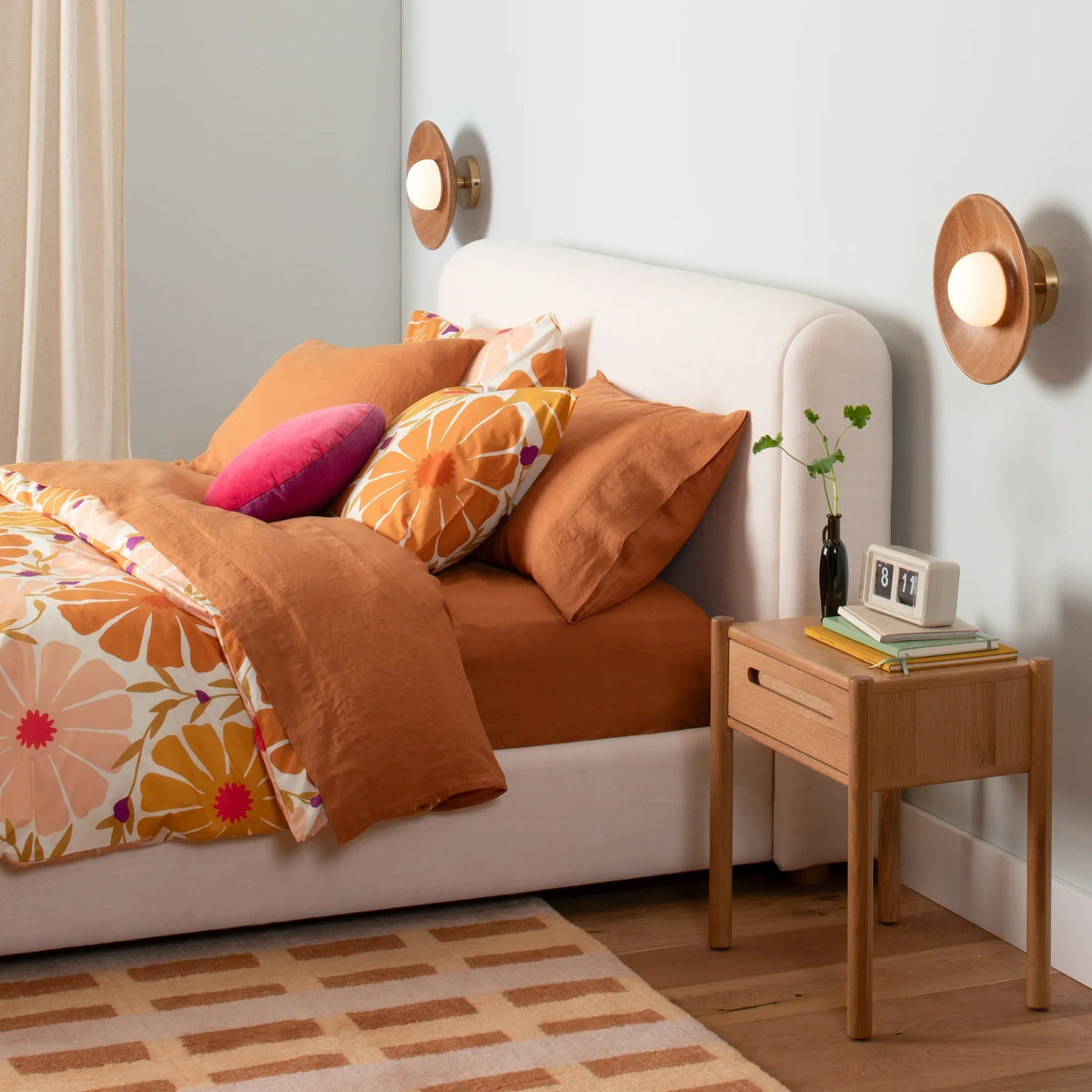 Bed with rust-orange duvet and floral cover, layered pillows and pink cushion beside an oak nightstand and wall sconces
