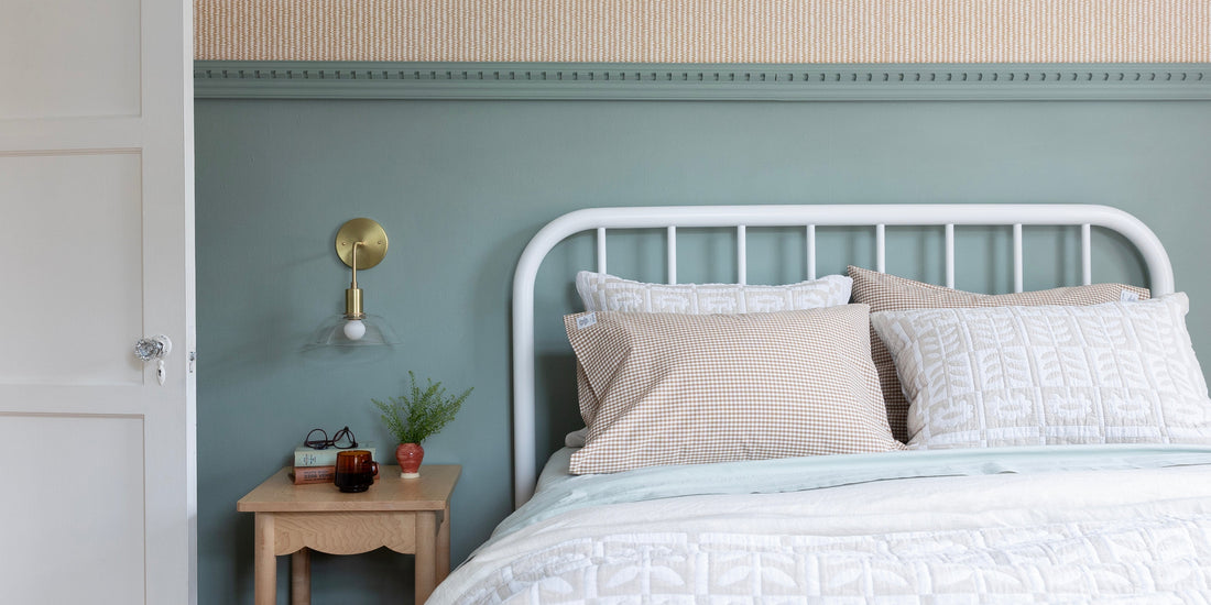 White metal bed with layered white quilt and gingham pillows beside a wooden nightstand and brass wall sconce against a soft green wall
