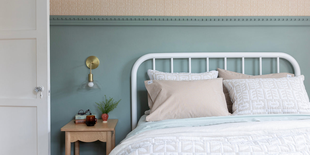 White metal bed with layered white quilt and gingham pillows beside a wooden nightstand and brass wall sconce against a soft green wall
