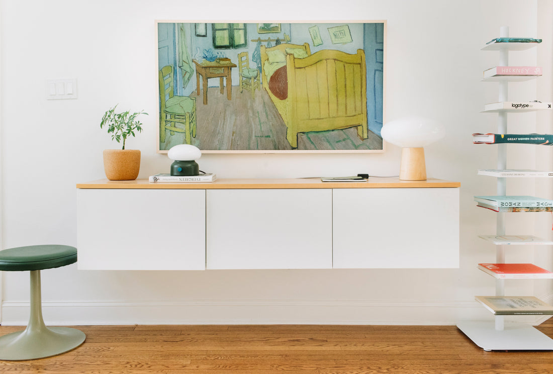 Floating white console displaying framed painting, potted plant, mushroom lamp, green stool, and narrow bookshelf in bright room