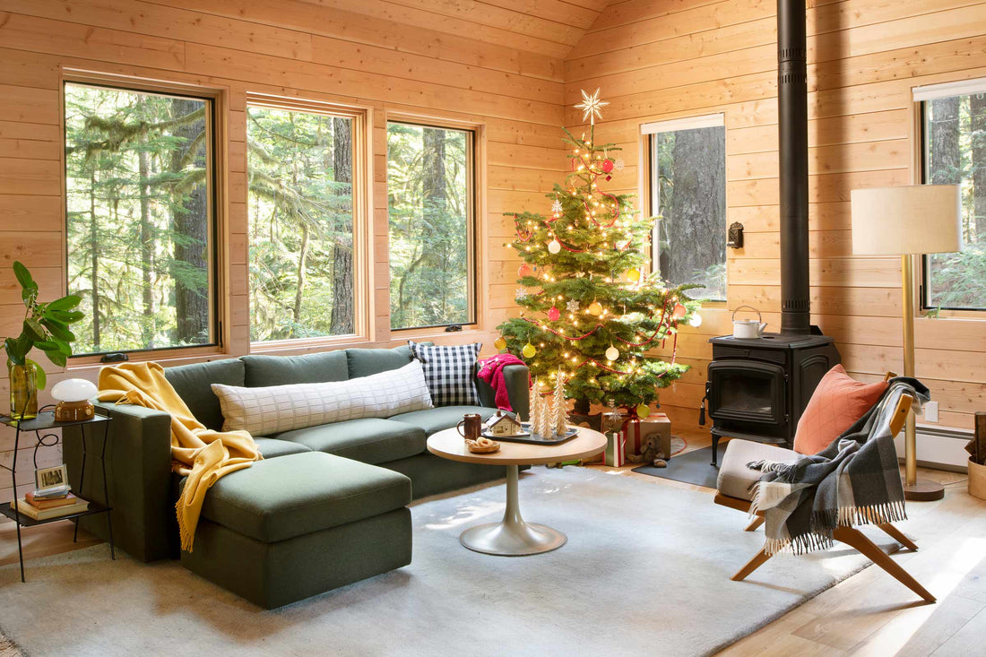 Cozy living room with decorated Christmas tree, green sectional sofa, wood stove, and windows overlooking an evergreen forest
