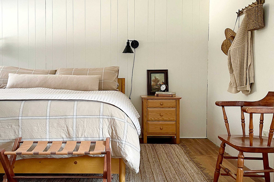 Cozy cabin bedroom with beige plaid bed, white paneled wall, wooden nightstand with framed art and alarm clock, woven rug