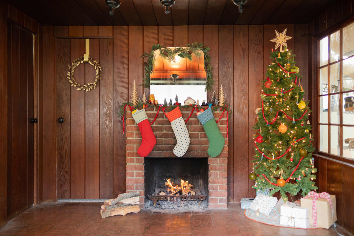 Three stockings hung on a brick fireplace ledge over a small fire, with a decorated Christmas tree and wrapped gifts to the right