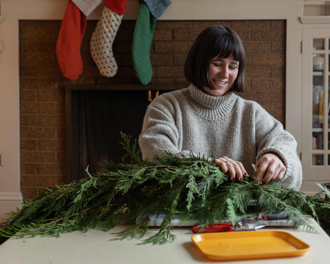 Person assembling an evergreen garland on a table with pruning shears, fireplace and stockings in the background