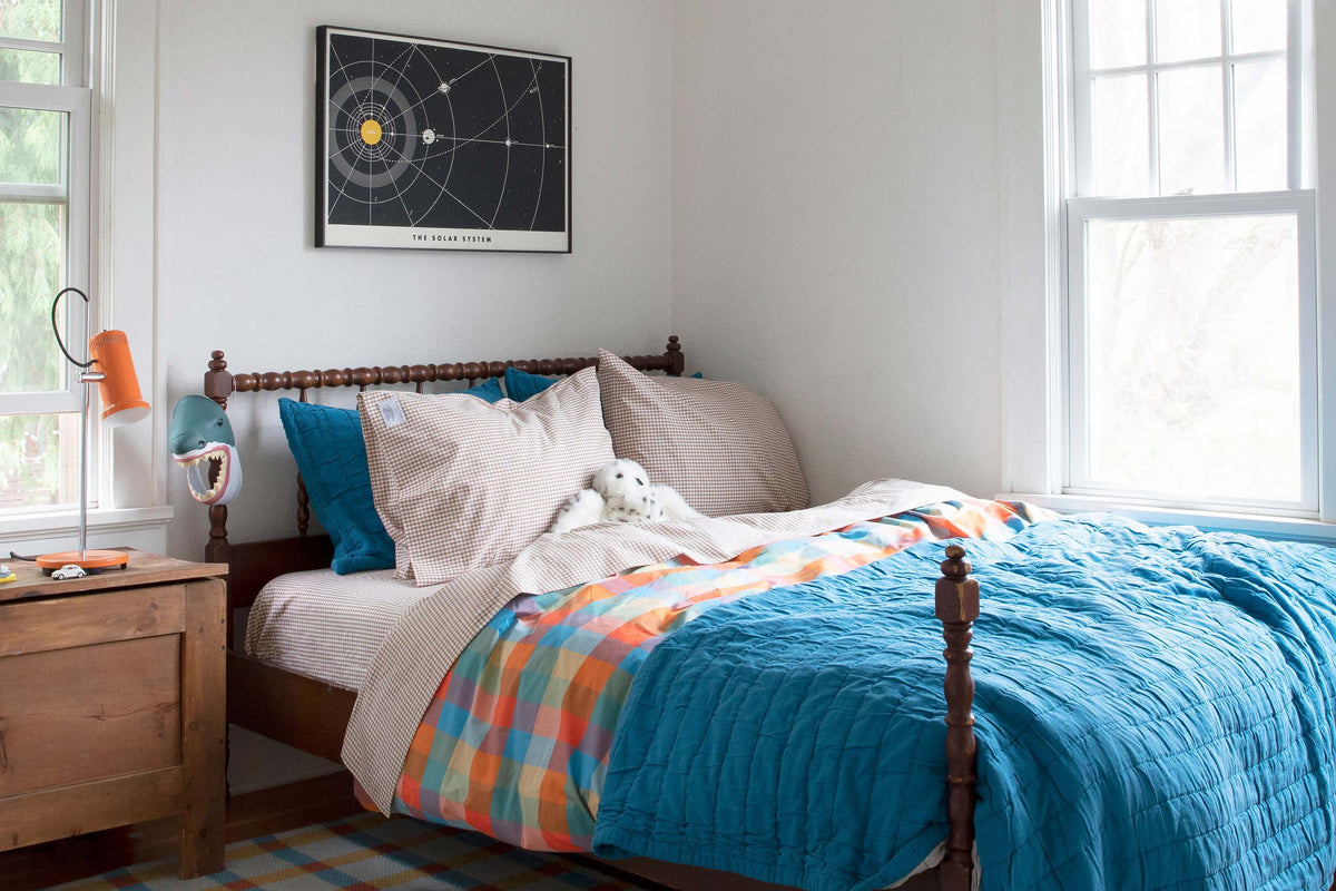 Sunlit children’s bedroom with wooden bed, bright blue quilt, multicolored plaid duvet, gingham pillows and plush seal