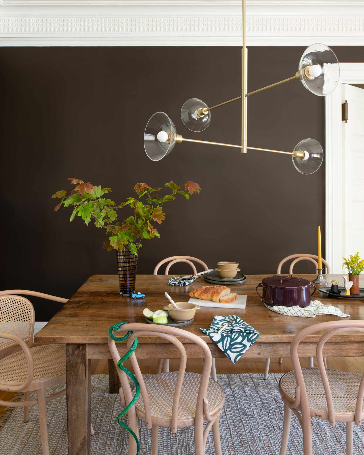 Dining table set for a casual meal beneath a brass multi-arm chandelier against a dark brown wall