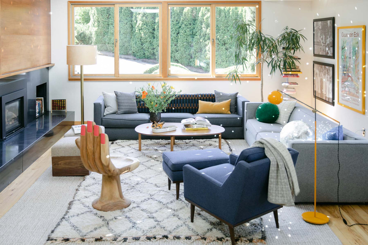 Sunlit living room with gray sectional sofa, blue armchair, wooden hand-shaped chair, patterned rug and coffee table with flowers by large window