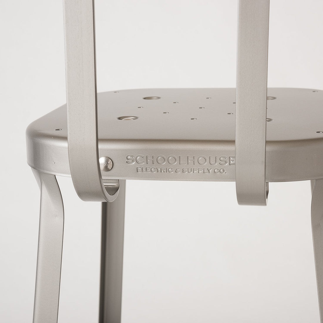 Close up of Utility Stool seat and back showcasing embossed SCHOOLHOUSE logo on raw coated steel with neutral background