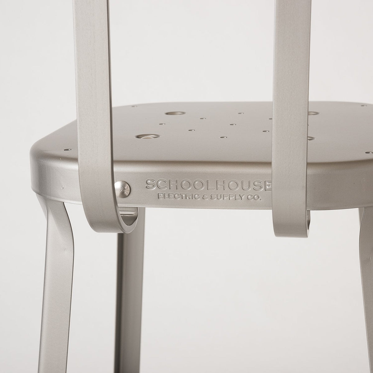 Close up of Utility Stool seat and back showcasing embossed SCHOOLHOUSE logo on raw coated steel with neutral background