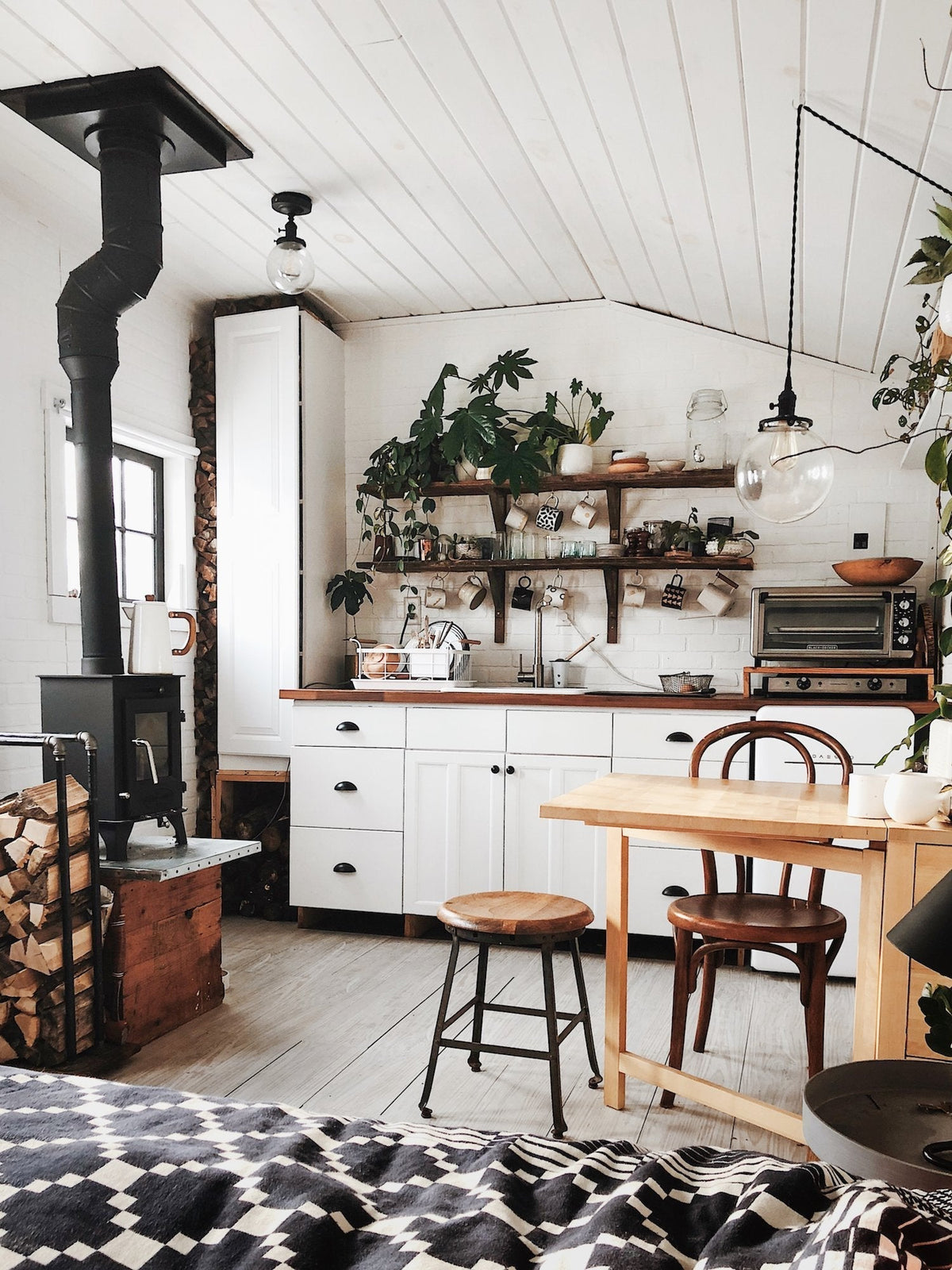 Cozy farmhouse kitchen with white cabinets, open wooden shelves holding plants and mugs, a wood stove, and a glass pendant light