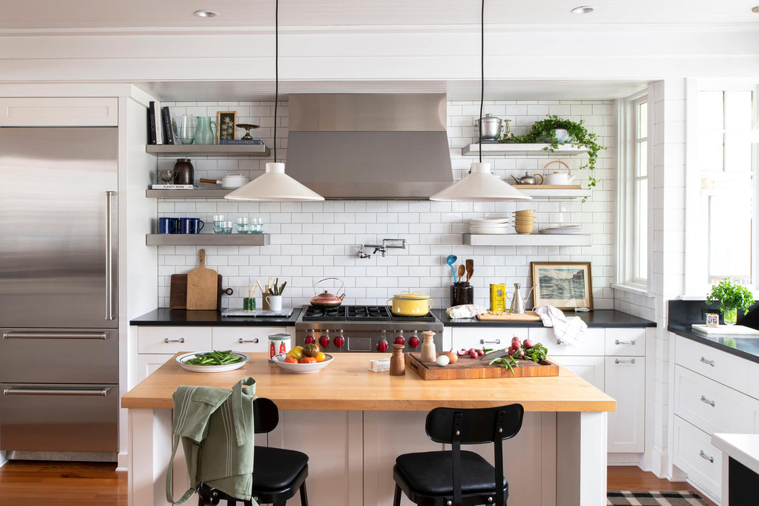 Two white pendant lights over a wood kitchen island with black stools, subway tile backsplash, stainless range, and open shelving