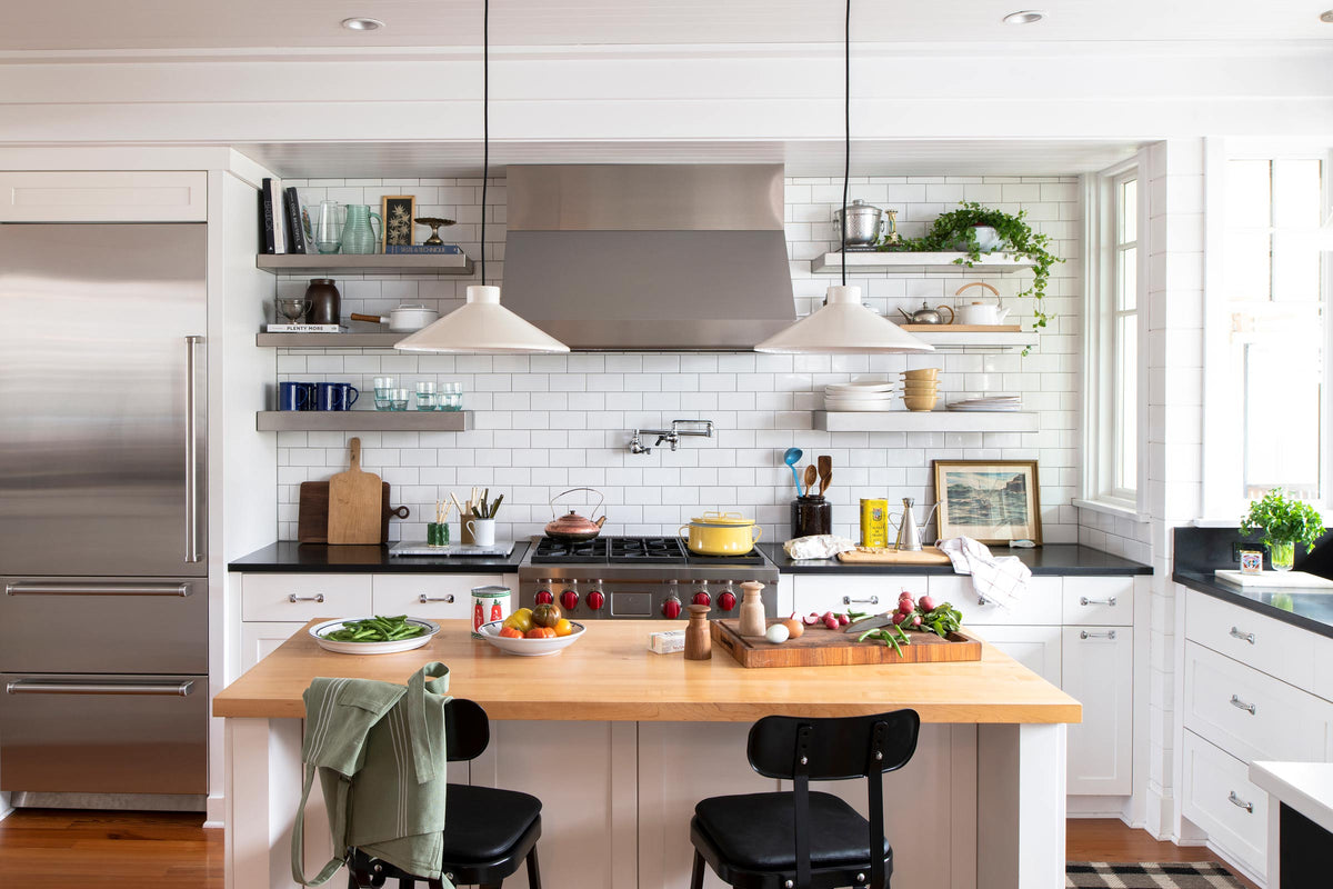Two white pendant lights over a wood kitchen island with black stools, subway tile backsplash, stainless range, and open shelving