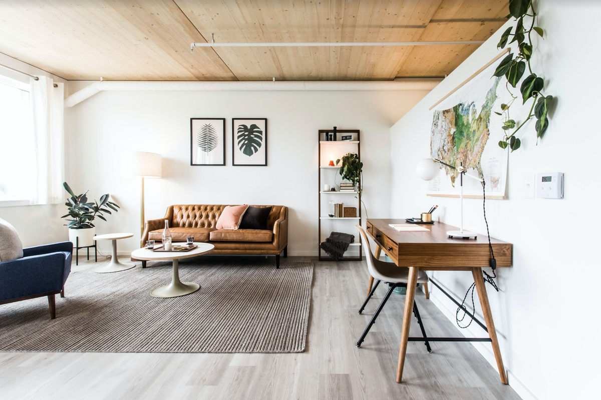 Sunlit open-plan living room featuring a tufted caramel leather sofa, round coffee table, mid-century wood desk, and wall map