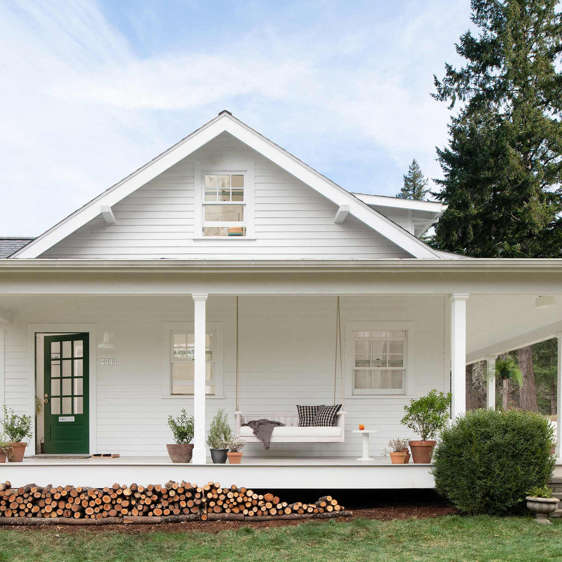 Front view of a white farmhouse-style home with a covered porch, green front door, hanging porch swing, potted plants, and stacked firewood under the porch