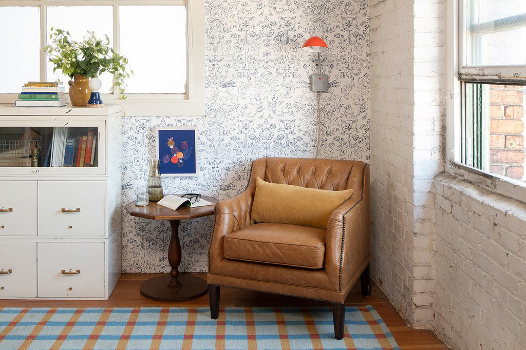 Leather tufted armchair beside a round wooden side table in a sunlit corner with floral wallpaper, white cabinet and plaid rug
