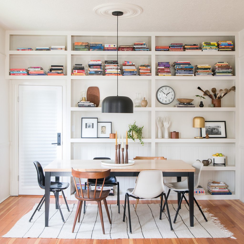 Dining table with mismatched chairs under a black pendant light set before white built-in bookshelves filled with colorful books