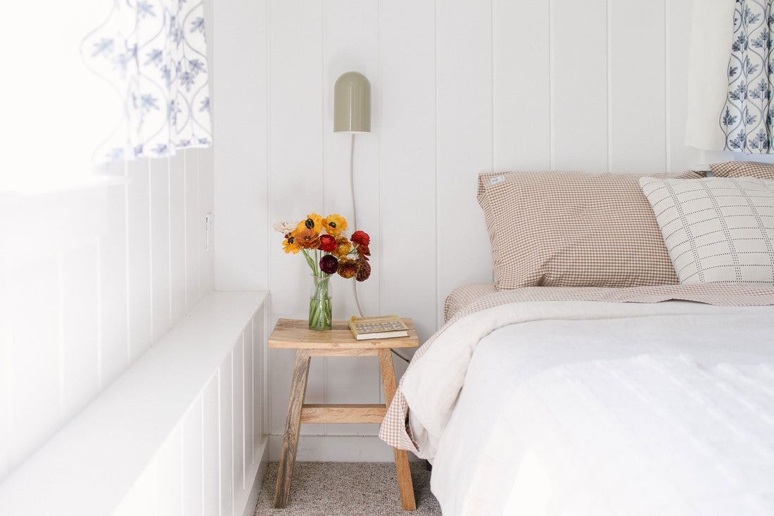 Bedside table with a vase of orange and red flowers next to a bed with beige gingham pillows and a green wall sconce in a bright white cottage bedroom