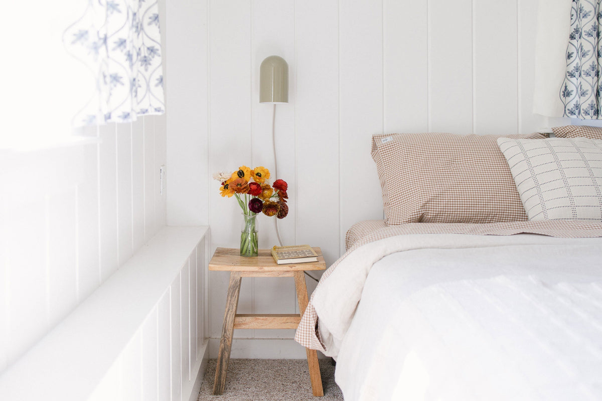 Bedside table with a vase of orange and red flowers next to a bed with beige gingham pillows and a green wall sconce in a bright white cottage bedroom