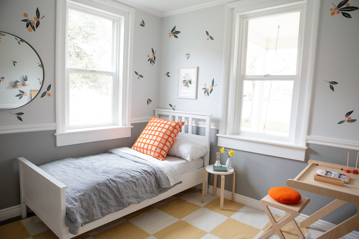 Child’s bedroom with white bed, gray bedding and orange check pillow, wall decals, windows, and wood desk with orange stool