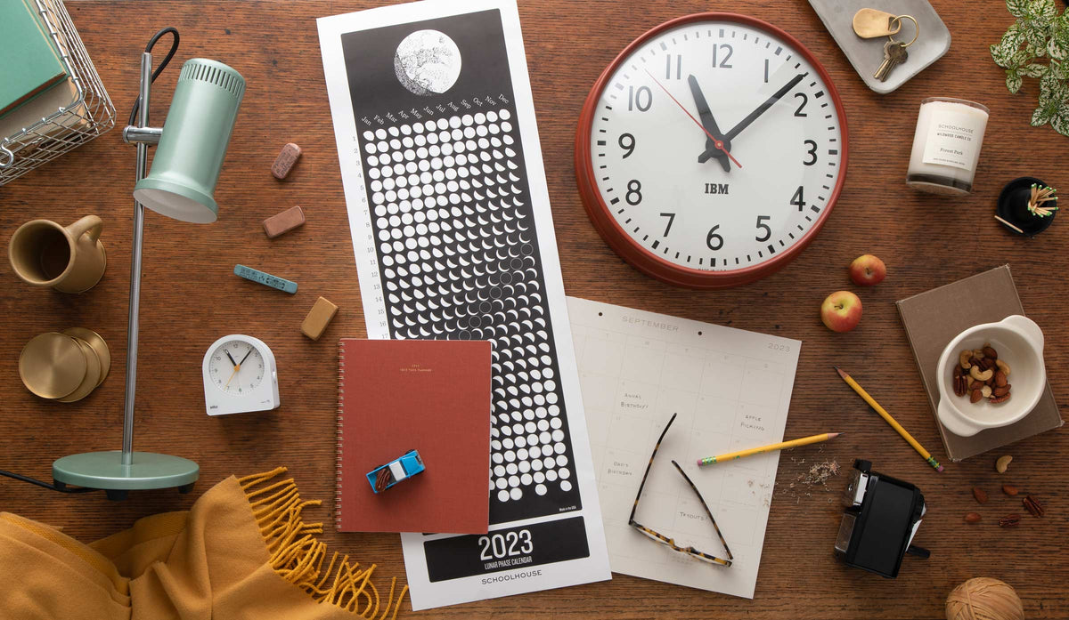 Overhead view of a wooden desk with a red wall clock, 2023 lunar calendar, green desk lamp, notebook, glasses, pencils, and snacks arranged as school supplies