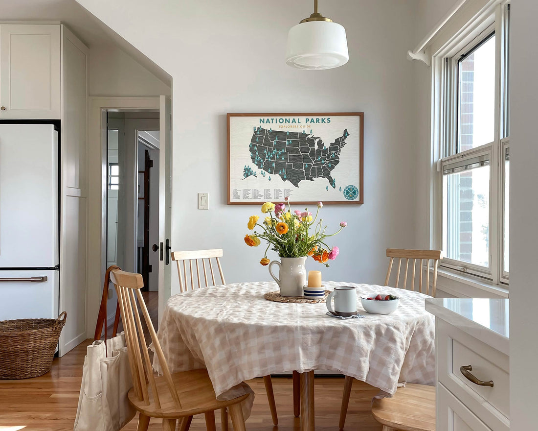Round dining table with gingham tablecloth and vase of flowers in a sunlit kitchen nook with a framed National Parks map