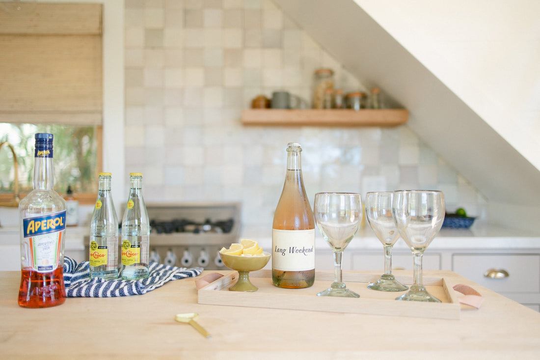 Kitchen counter set for Aperol spritz with Aperol bottle, Long Weekend wine, lemon wedges and three wine glasses on tray