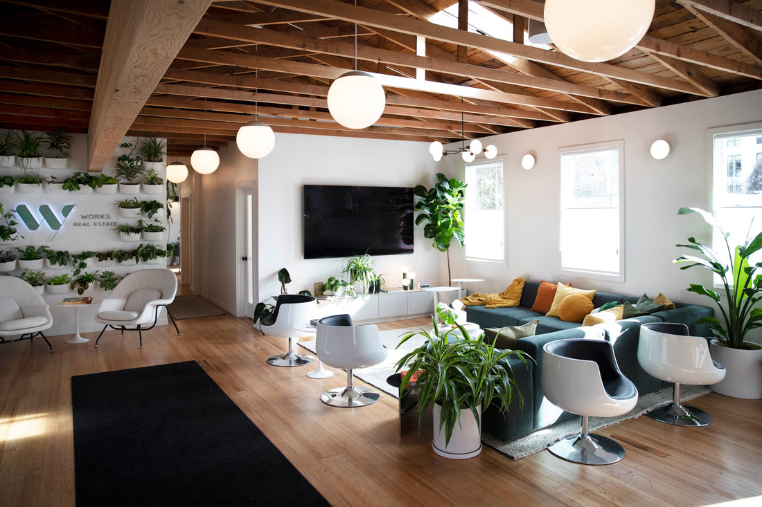 Bright plant-filled office lounge with a green sectional, white swivel chairs, exposed wood ceiling beams, globe pendant lights, and a living plant wall